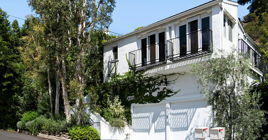 exterior shot of tall white LA home featuring trees, vines, two garage doors, and balconies. 