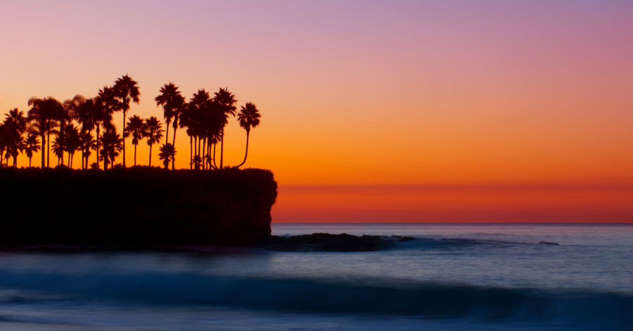 A bright red, yellow, and orange sunset with palm tree silhouette over dark blue ocean waters at Crescent Bay Beach