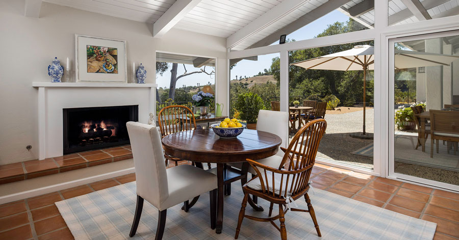 Kitchen table with a large fireplace and while mantel plus a glass wall of windows and doors that lead to the backyard. 