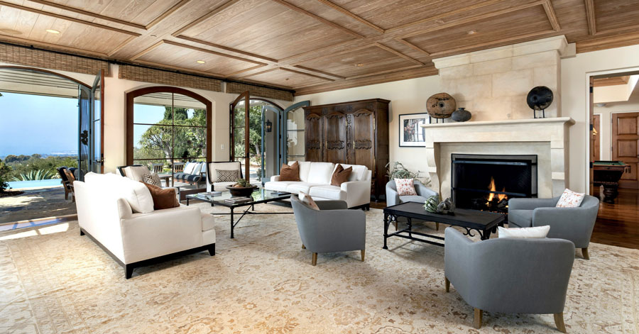 Large room with wood paneled ceilings, a lush white and tan rug, and seating area in front of a white stone fireplace. 