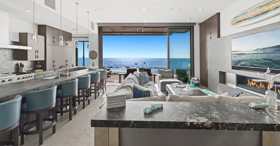 Kitchen and living area that looks directly out at blue ocean water with a wide but short fireplace in front of a large couch. 