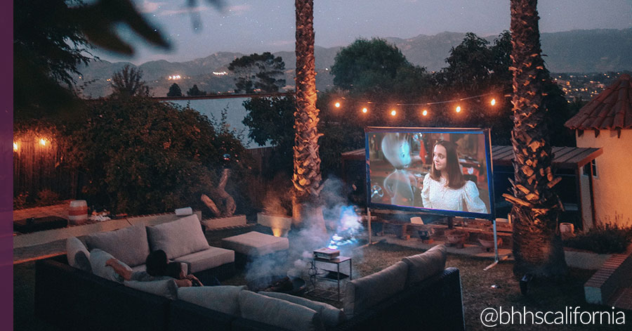 Outdoor home theater with couch, projector, screen, fire pit surrounded by lights and palm trees. 