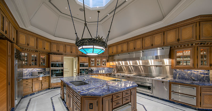 Large kitchen featuring blue French marble counter tops and brown wood cabinets. Large colored glass chandelier is suspended from ceiling with sky light above