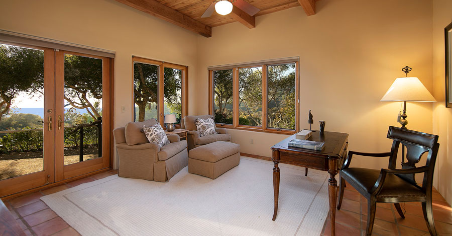 Home office with brown beamed ceilings, beige walls, and brown trim. French doors open to a peaceful outdoor backyard