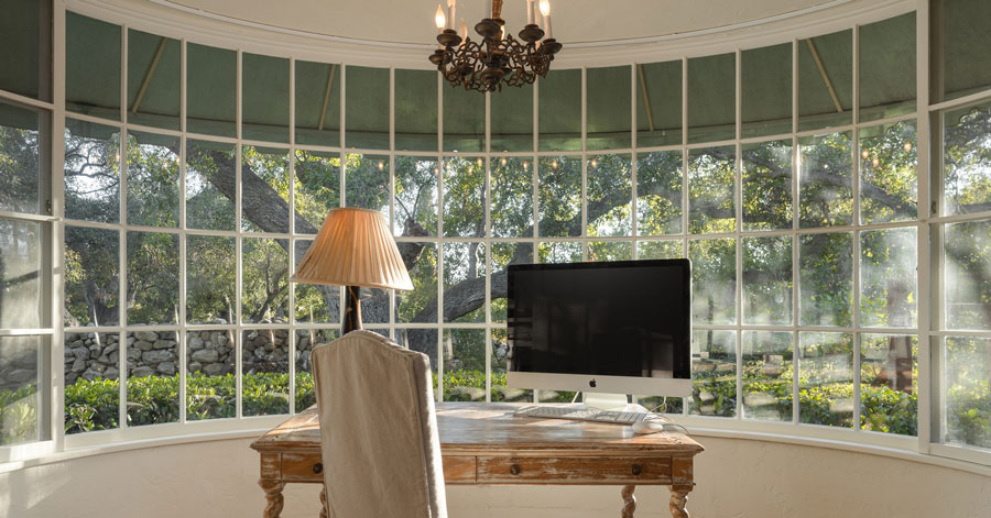 Home office with large curved picture window that looks out at peaceful backyard, tree, and green foliage 