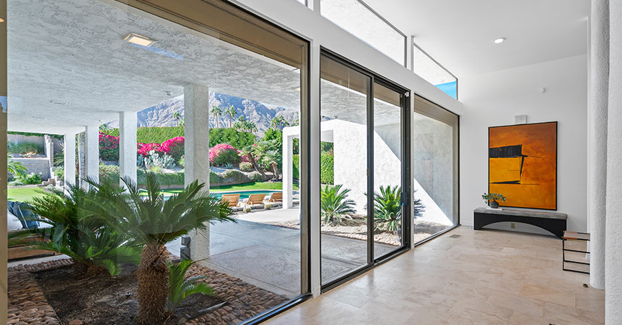 Interior of Mid-century modern home with floor to ceiling glass walls and doors looking out on pool, palm trees, and mountains