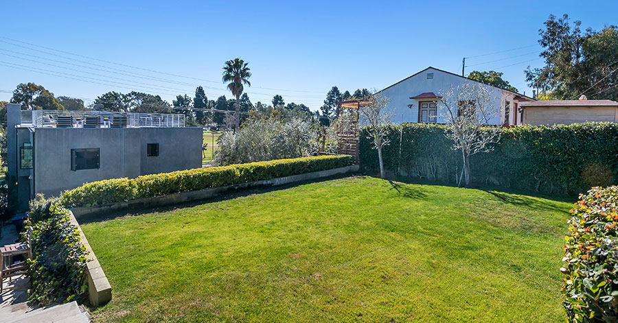 Large grassy area with two trees and bushes lining the edges with homes nearby and stone steps leading down 