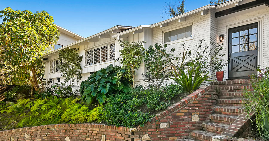 exterior of home with white brick walls, red brick steps and retaining wall, and black front door surrounded by lush green plants 